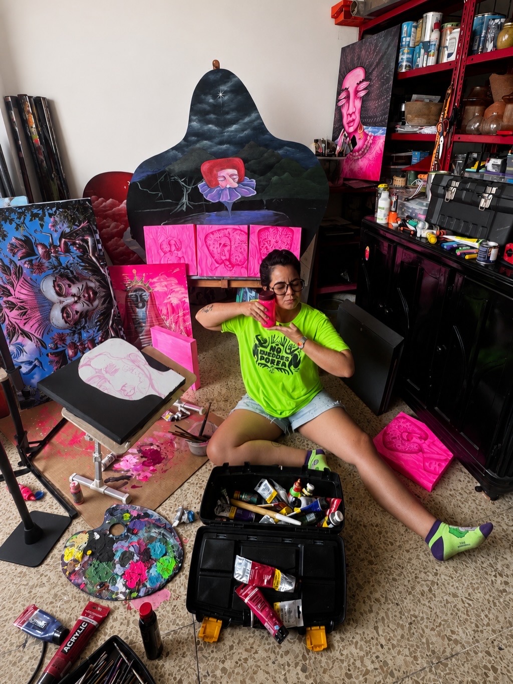 Carla Bresciani sentada en el piso de su taller, rodeada de pinturas en proceso en rosa fluorescente y una pieza recortada con forma de manto